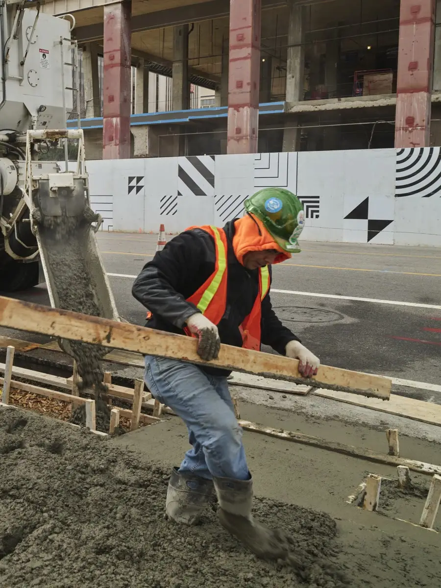 Un trabajador con casco verde y chaleco naranja empareja concreto recién vaciado en una obra