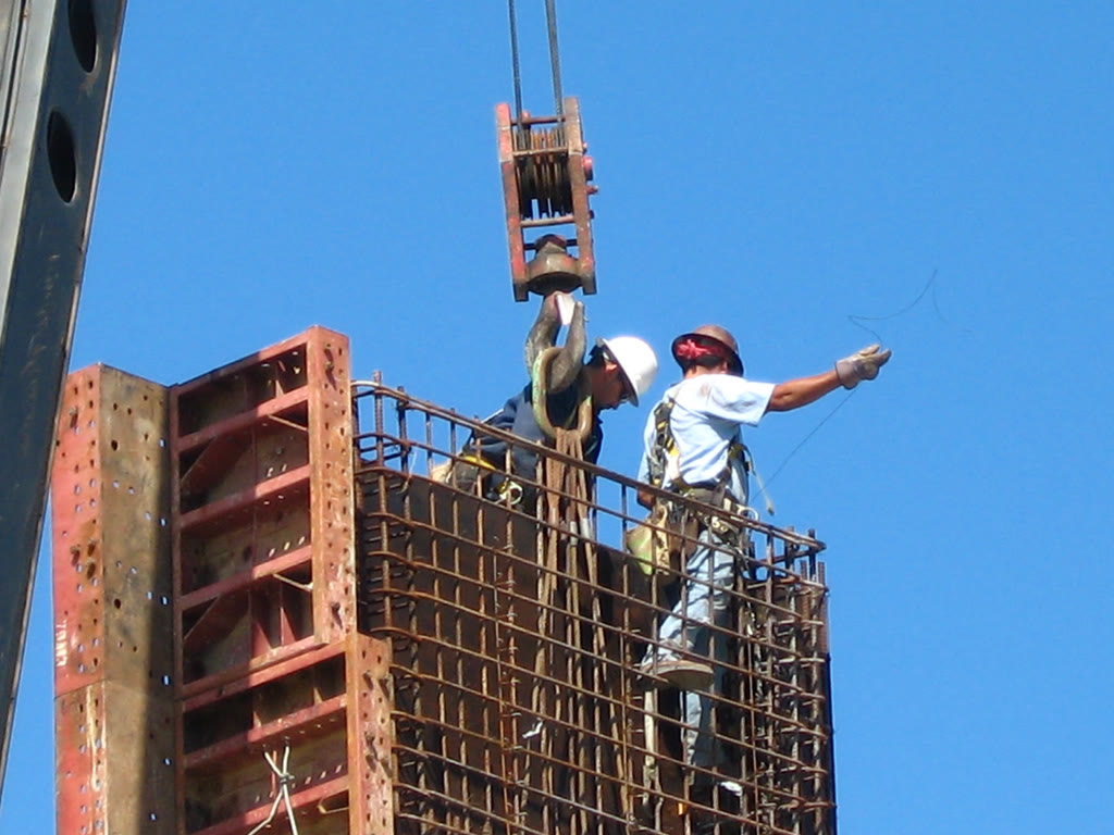 Dos trabajadores con casco arman una jaula de acero de refuerzo que cuelga de una grúa, contra el cielo azul